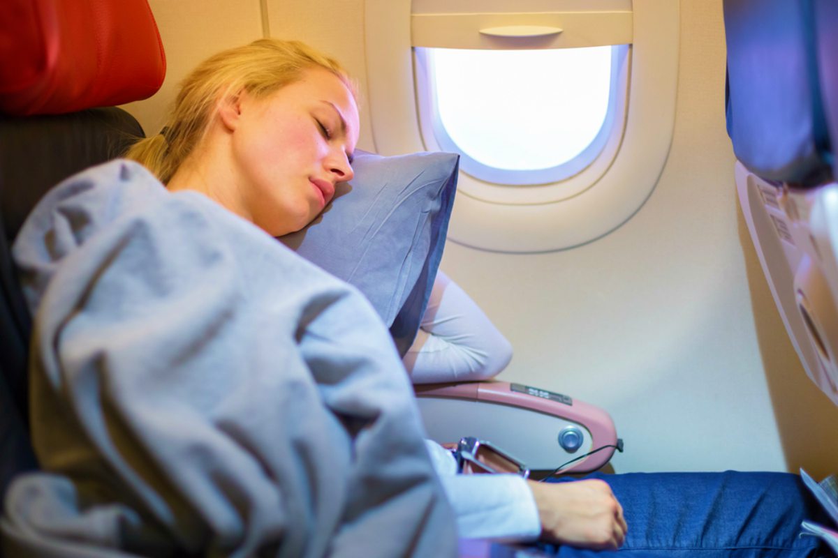 Tired blonde casual caucasian lady napping on uncomfortable seat while traveling by airplane | Travel Essentials For A Healthy Holiday