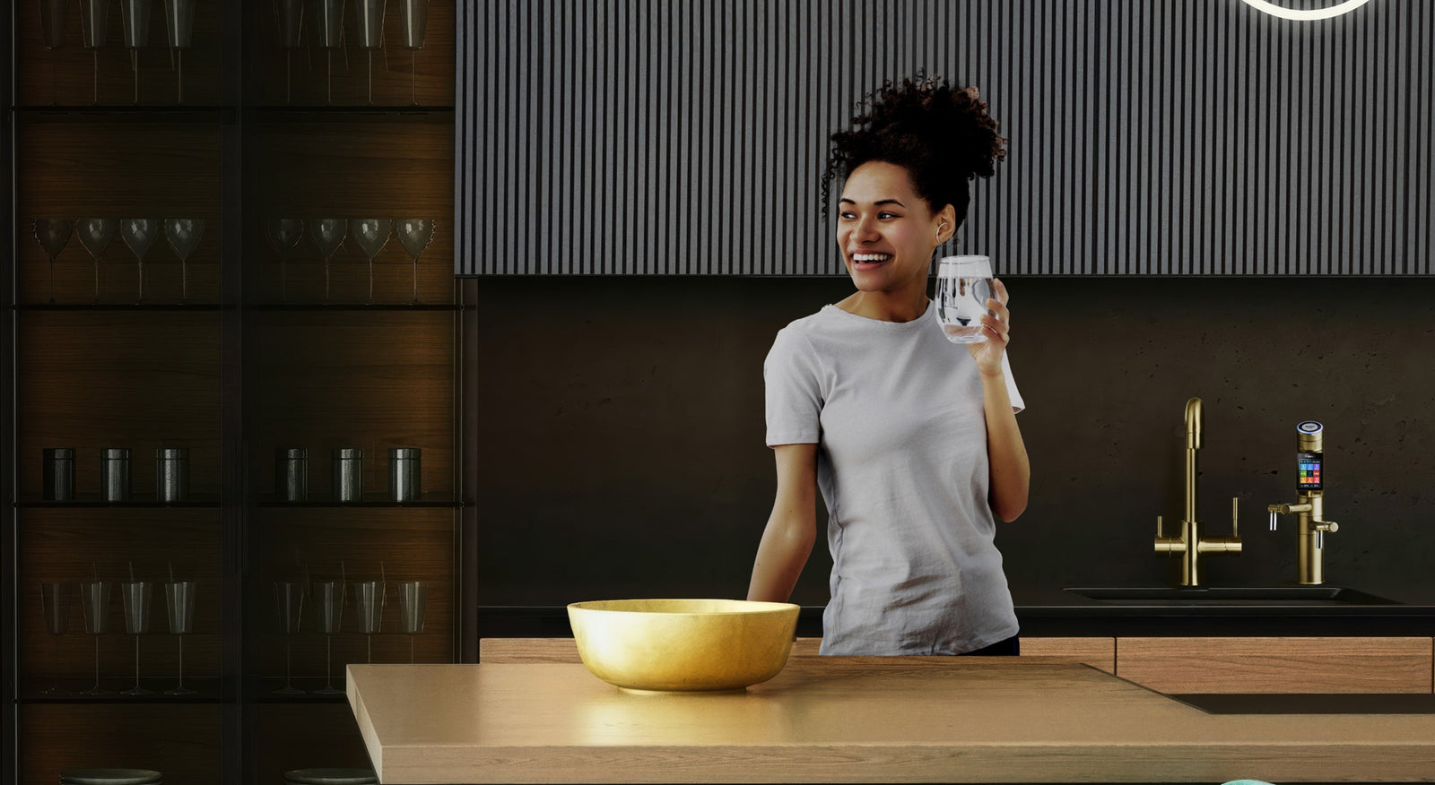 A woman smiling holding a glass of water in a luxurious modern kitchen with a Tyent UCE gold water ionizer in the background.